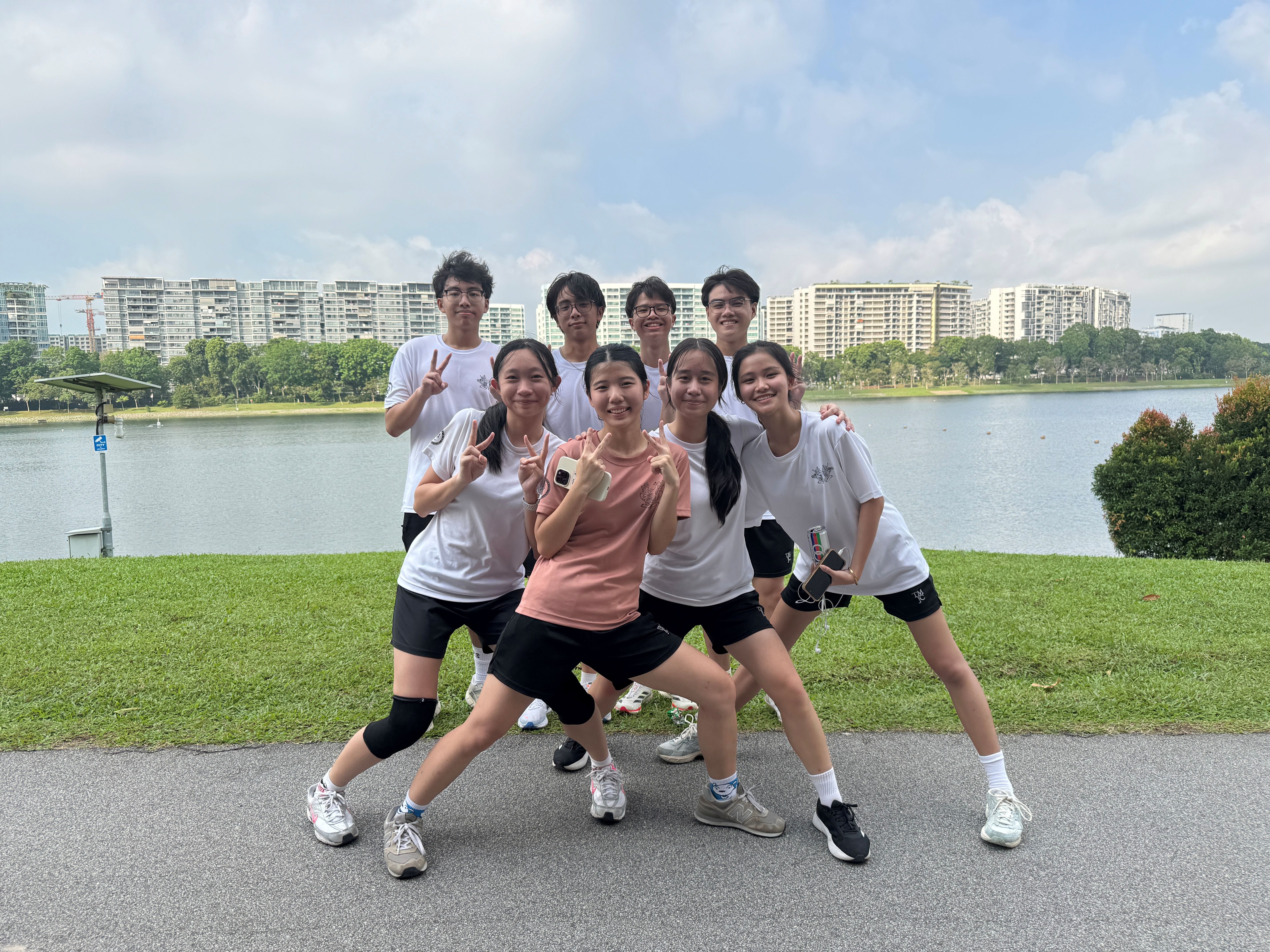 Eight students in white T-shirts and black shorts pose cheerfully by a reservoir, with four boys standing behind four girls who strike fun poses.